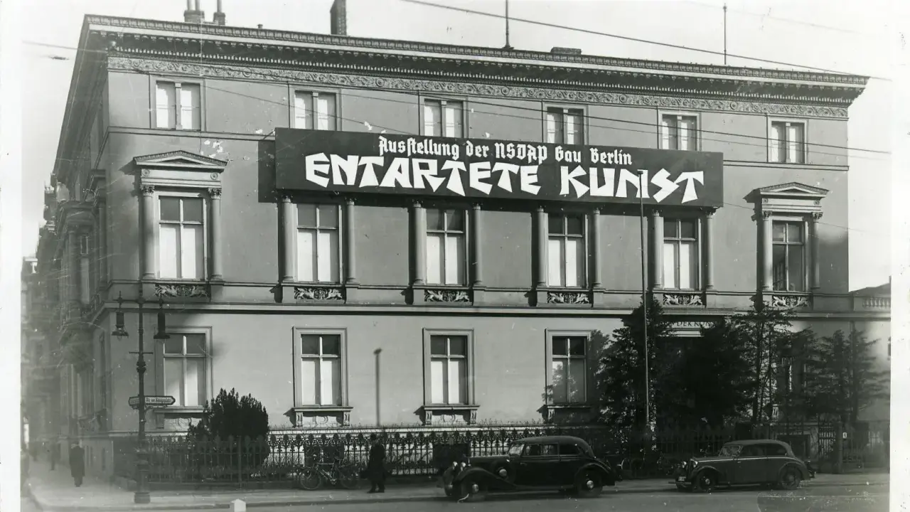 Große Banner verkünden die "Ausstellung der NSDAP Gau Berlin ENTTARTETE KUNST" an einem Gebäude. Autos und Fahrräder stehen davor.