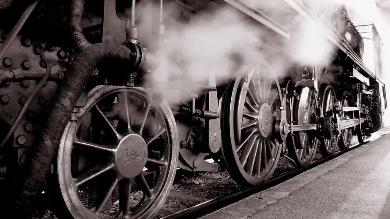 Dampf steigt auf von den Rädern einer historischen Lokomotive, ein Symbol für die Erfindung der Eisenbahn und den Beginn einer neuen Ära.