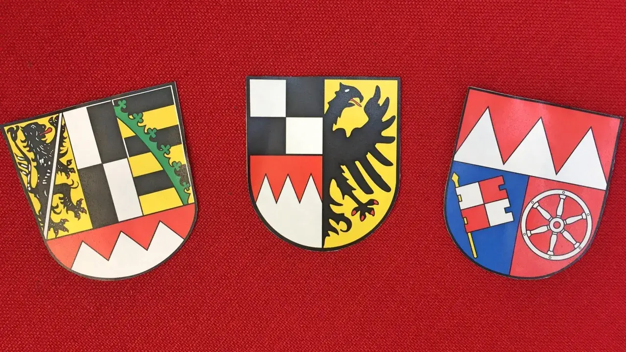 Drei Wappen auf rotem Grund: links ein Löwe, Mitte ein Adler, rechts ein Fähnlein und ein Rad. Sie repräsentieren bayerische Regierungsbezirke.