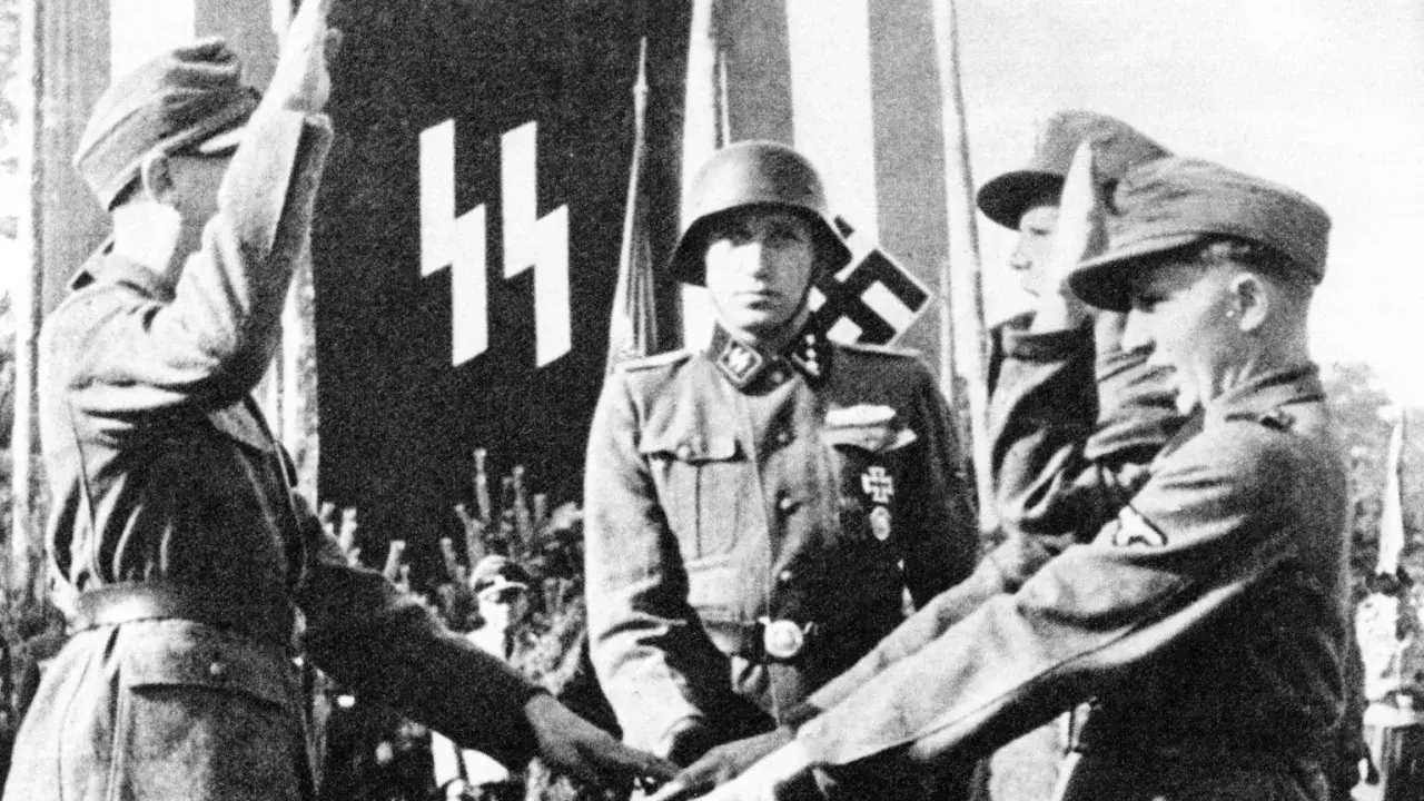Mehrere SS-Soldaten stehen vor einer Fahne mit SS-Runen. Einer hebt den Arm zum Gruß.