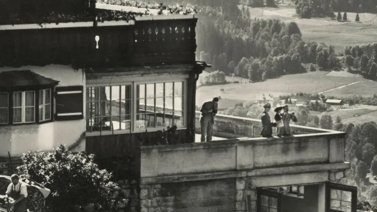 Blick auf die Hitler-Residenz mit Personen auf dem Balkon und einer malerischen Landschaft im Hintergrund.