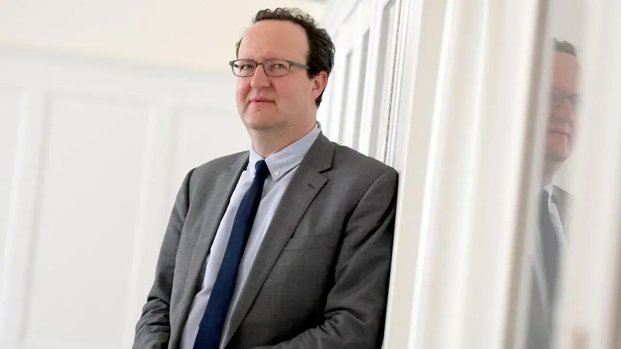 Hans Ottomeyer, ein Mann mit Brille im grauen Anzug, lehnt an einer weißen Wand.