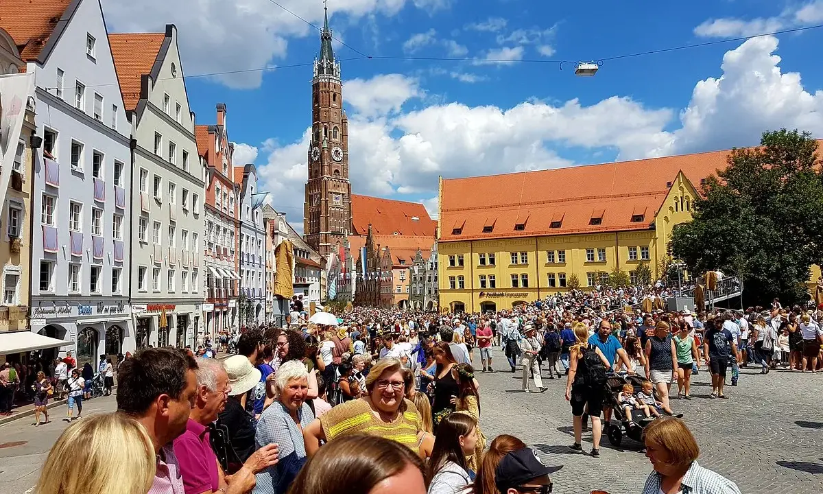 Menschenmassen auf dem Platz der Landshuter Hochzeit, mit historischen Gebäuden und einem Kirchturm im Hintergrund.