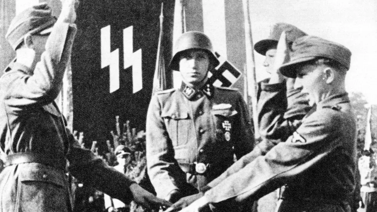 Ein SS-Offizier im Helm steht vor einer Fahne mit SS-Runen. Mehrere junge Männer in Uniformen sind zu sehen.