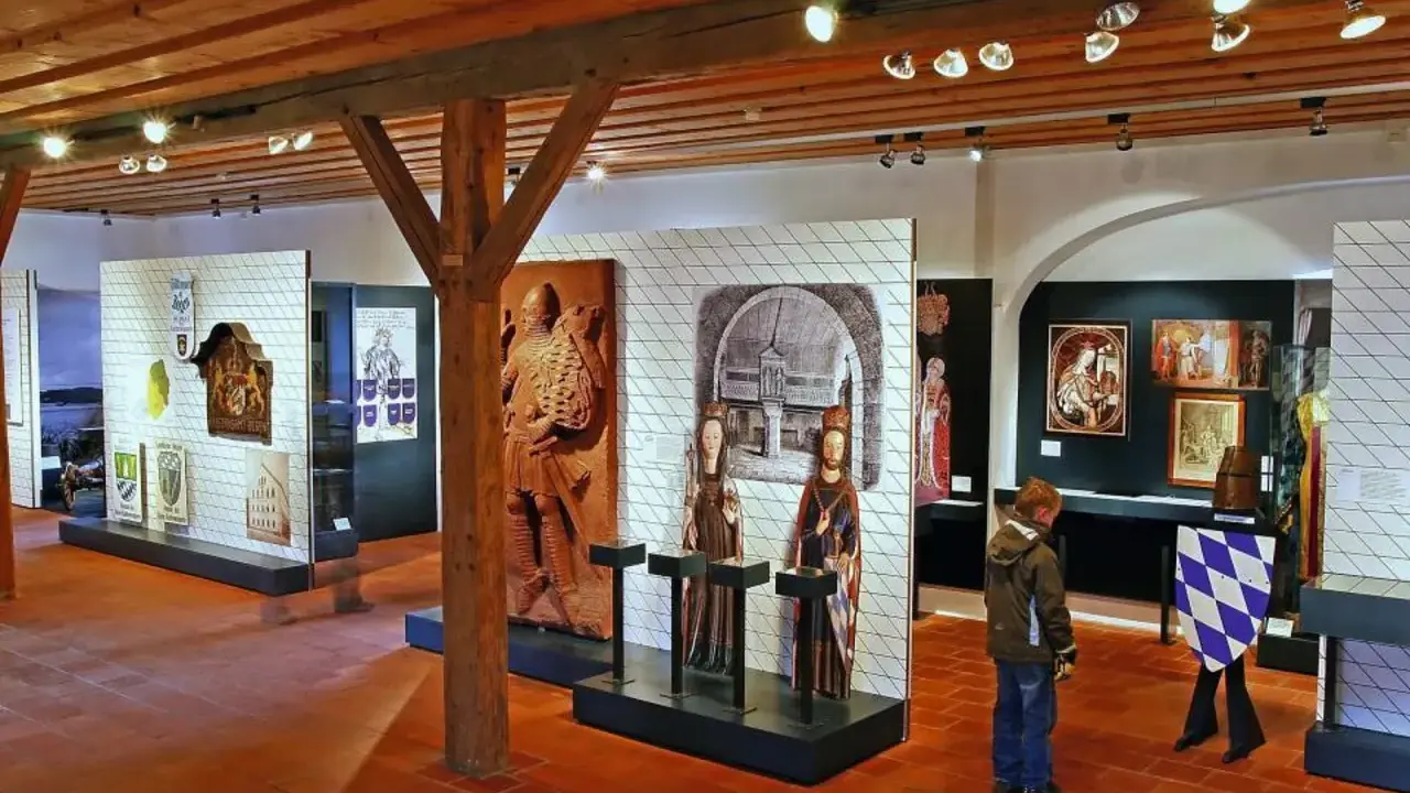 Museumsausstellung mit historischen Artefakten, darunter das Wittelsbacher Wappen, Skulpturen und Gemälde.