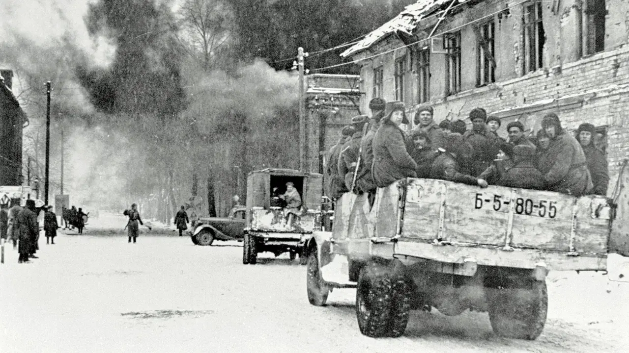 Soldaten der Roten Armee 1945 fahren in einem LKW durch eine verschneite Stadt. Rauch steigt aus einem zerstörten Gebäude auf.