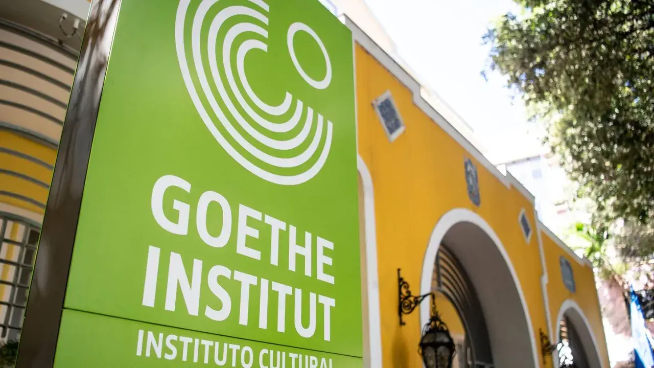 Grünes Schild mit weißem Logo und Schriftzug "GOETHE INSTITUT" vor einem gelben Gebäude.
