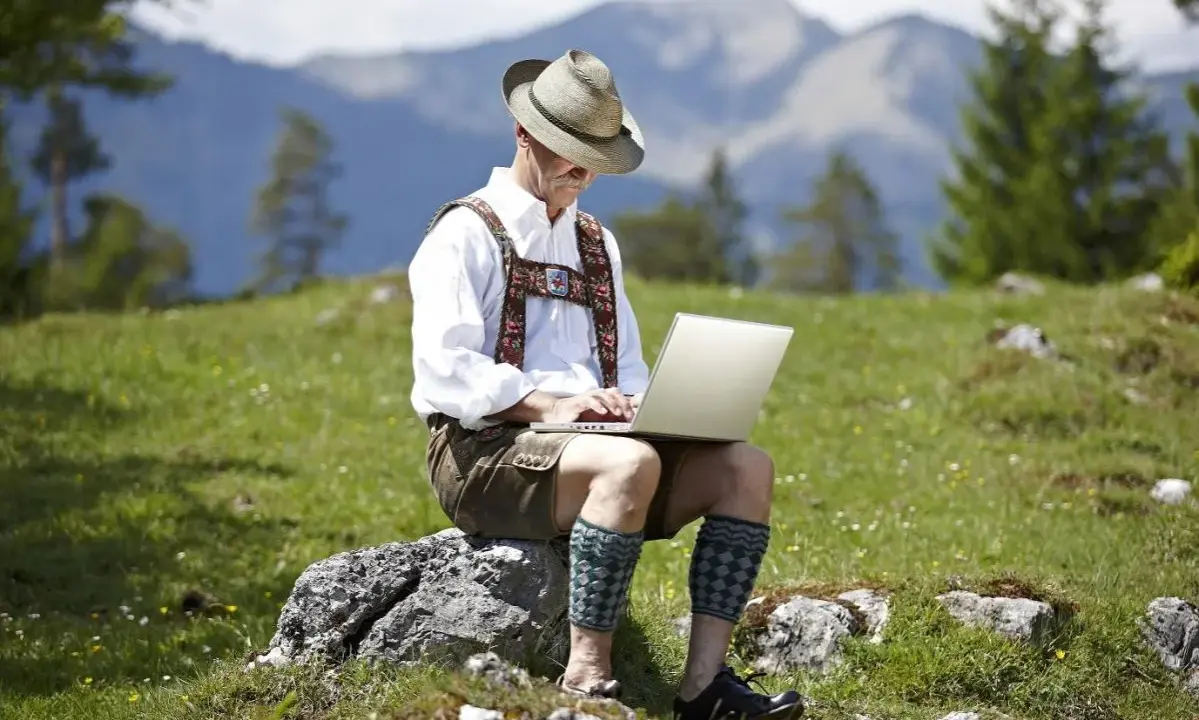 Mann in Lederhose und Hut arbeitet auf einem Laptop in den Bergen.