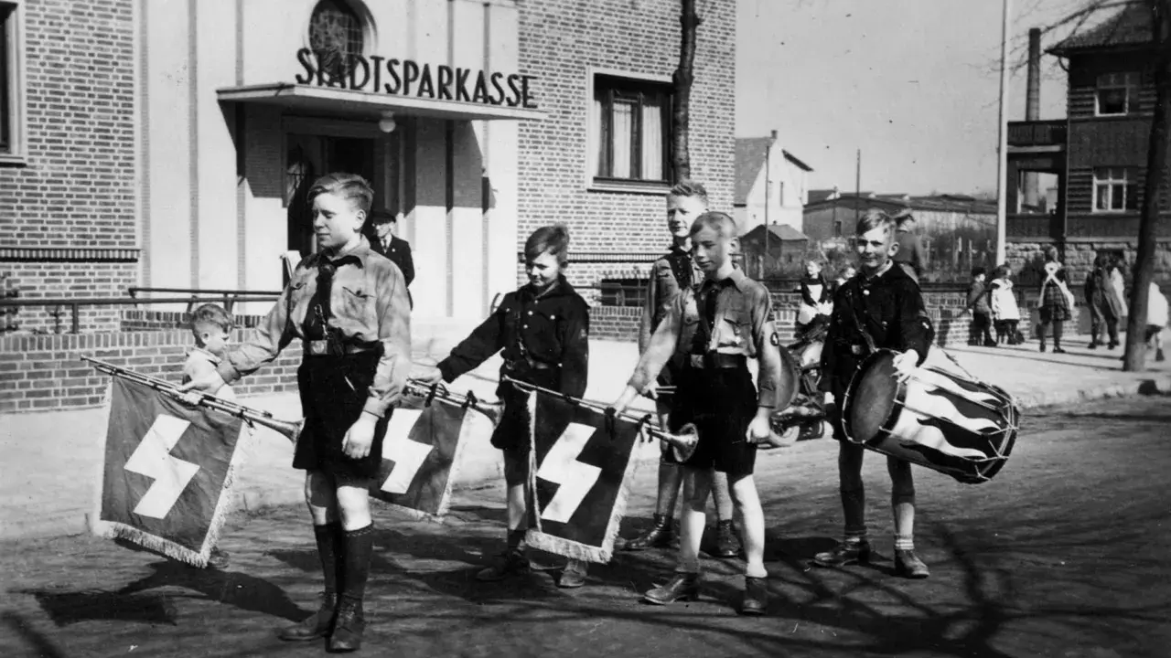 Jungen in Uniformen tragen Fahnen mit Hakenkreuzen und Trommeln. Ein junger Mann, der an Hitler erinnert, marschiert mit ihnen.