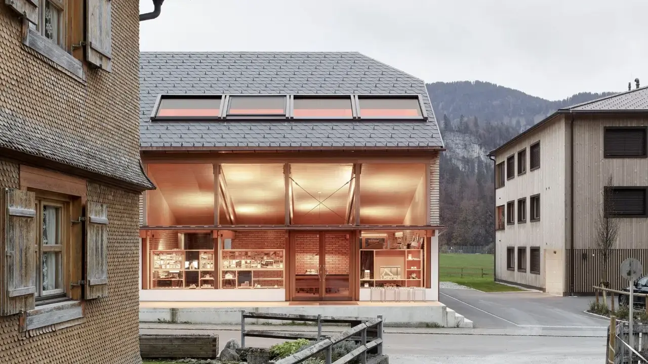Neues Bauen: Moderne Architektur trifft auf traditionelle Schindelfassade. Ein hell erleuchtetes Glasgebäude mit Ausstellungsstücken steht neben einem Holzhaus.