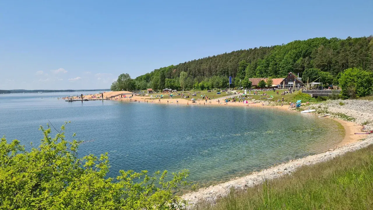 Sonniger Strandtag am Rothsee bei Heideck, Bayern. Menschen entspannen sich am Sandstrand, während Segelboote auf dem Wasser gleiten.