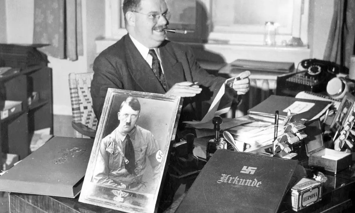 Philipp Auerbach sitzt an seinem Schreibtisch, eine Pfeife im Mund, und betrachtet ein Bild von Adolf Hitler. Daneben liegt eine Urkunde.
