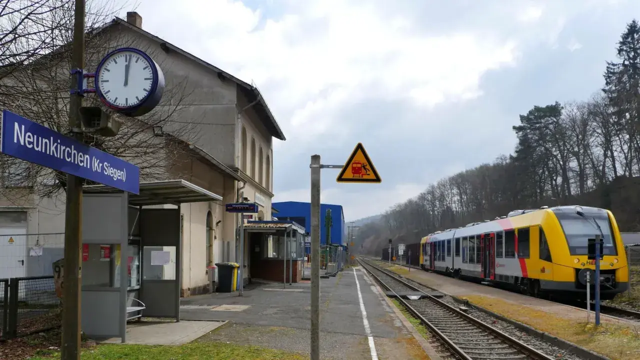 Bahnhof Neunkirchen (Kr Siegen) mit gelb-silbernem Zug auf den Gleisen. Ein Warnschild zeigt einen Zug und eine stürzende Person.