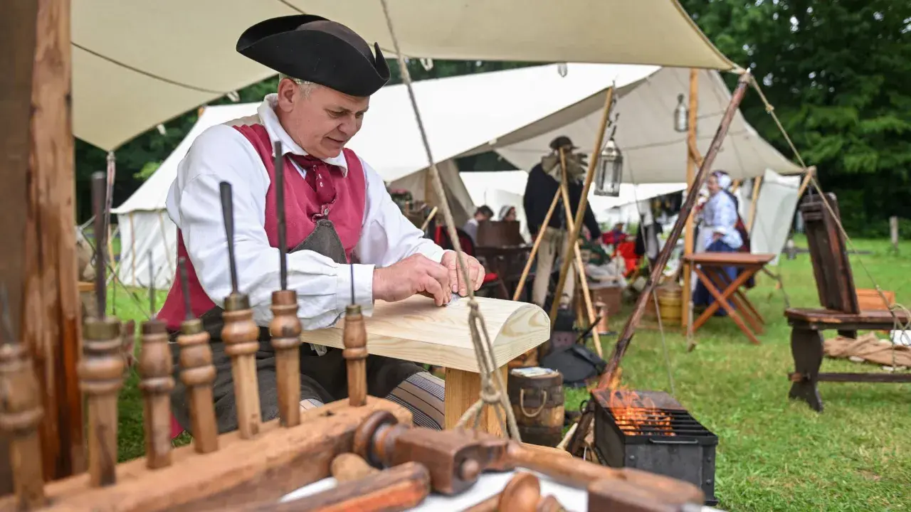 Ein Handwerker in historischer Kleidung arbeitet an Holz. Dies ist ein Beispiel dafür, was ist ein Freilichtmuseum: lebendige Geschichte zum Anfassen.