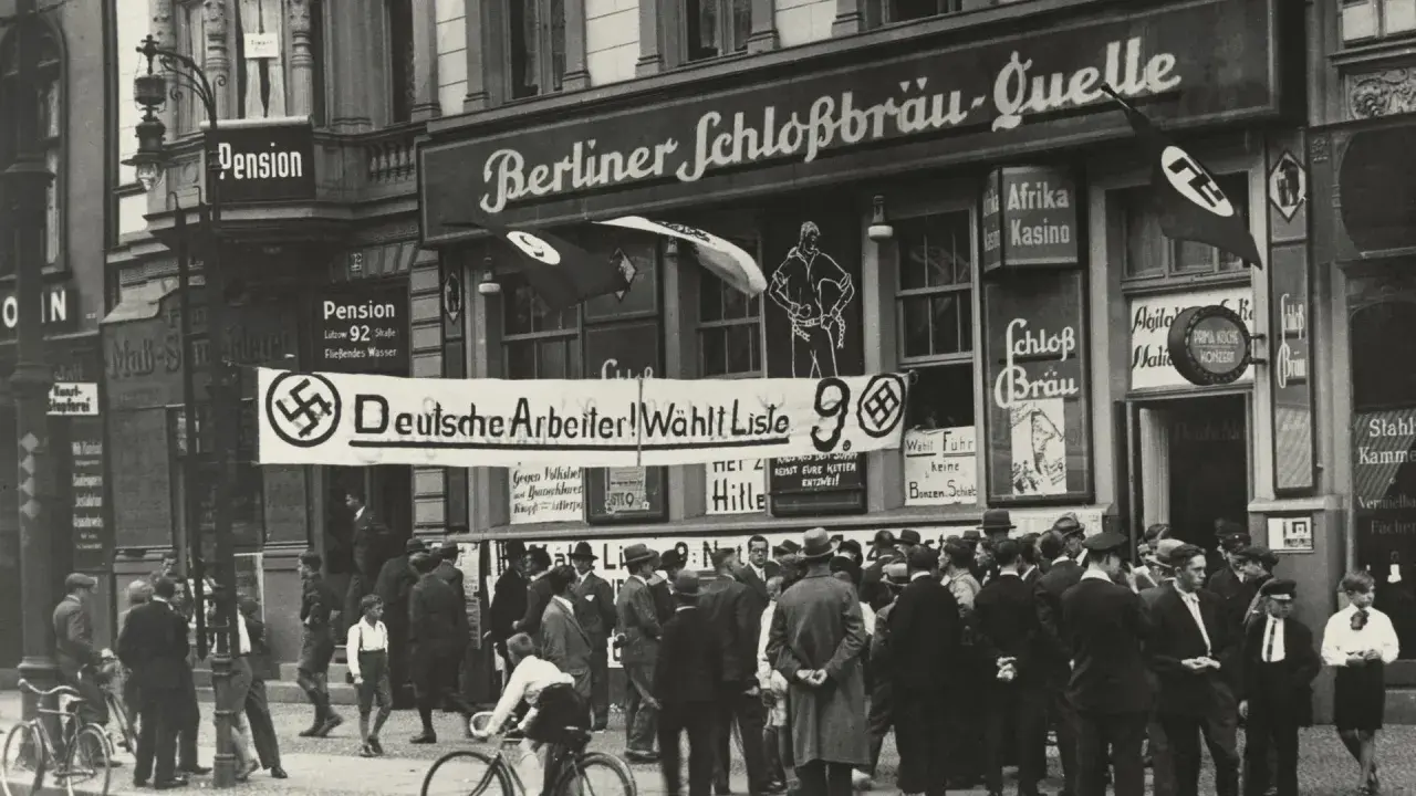 Menschenmenge vor einem Gebäude mit der Aufschrift "Berliner Schloßbräu-Quelle". Ein Banner verkündet "Deutsche Arbeiter! Wählt Liste 9". Hakenkreuzfahnen sind sichtbar, was auf die Zeit vor dem NSDAP Verbot hinweist.