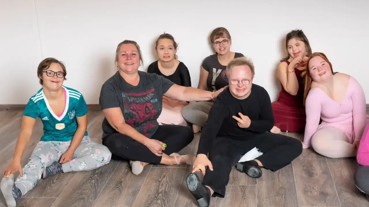 Gruppe von Menschen mit Down-Syndrom und einer Betreuerin sitzen auf dem Boden.