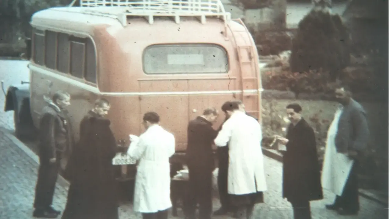 Männer in weißen Kitteln und dunklen Mänteln stehen vor einem roten Bus. Ein Kennzeichen ist sichtbar. Die Szene könnte mit der NS-Euthanasie in Verbindung stehen.