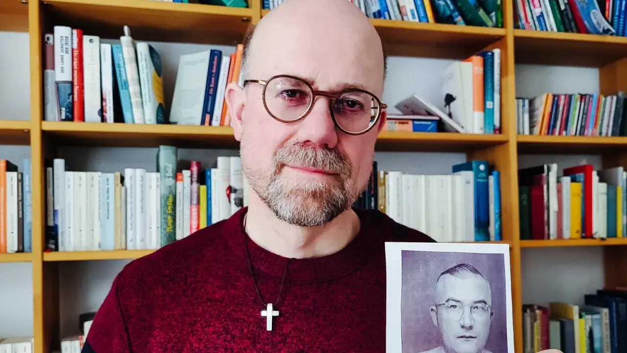 Mann mit Brille hält Foto eines Mannes in Uniform. Im Hintergrund Bücherregale. Das Foto zeigt einen Mann, der der SS Schutzstaffel angehörte.