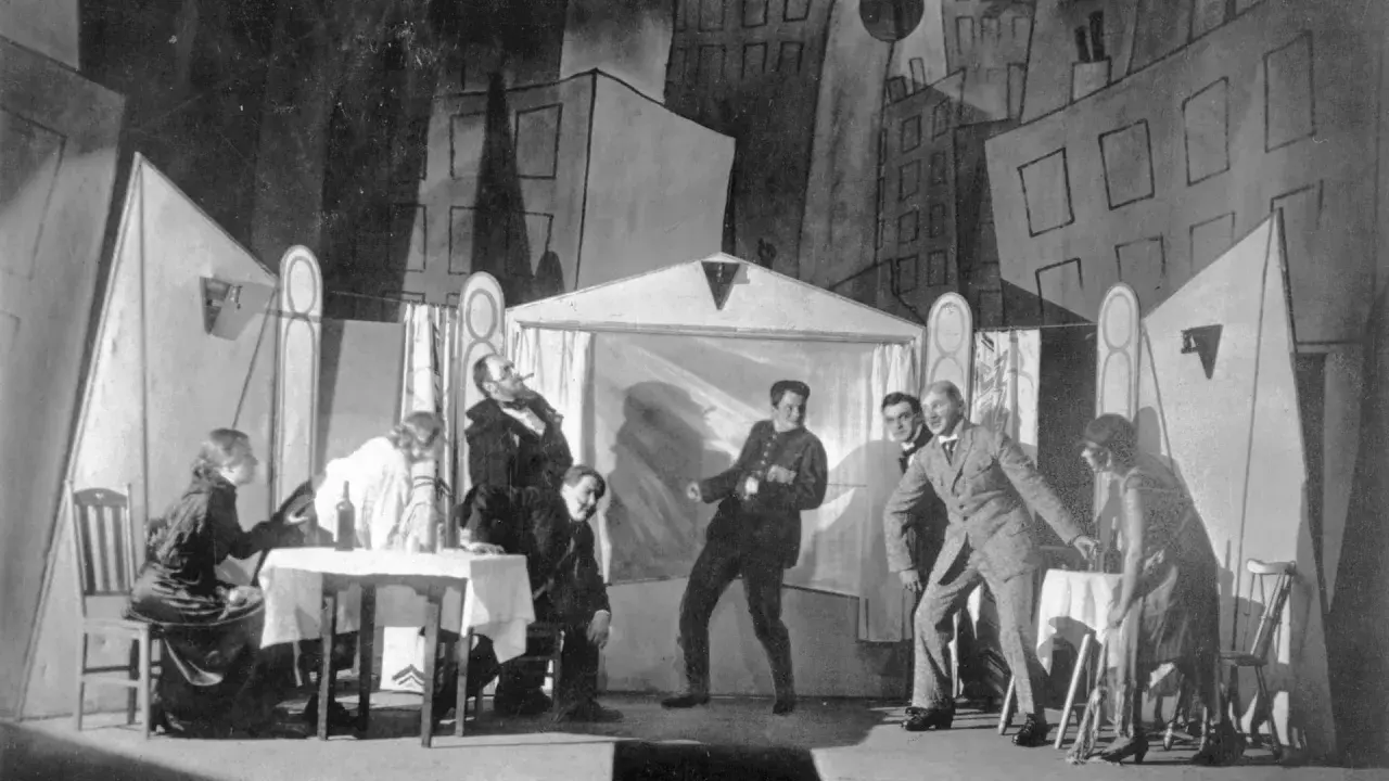Szene einer Theateraufführung mit expressionistischem Bühnenbild. Menschen agieren auf der Bühne, als würden sie **trommeln in der Nacht**.