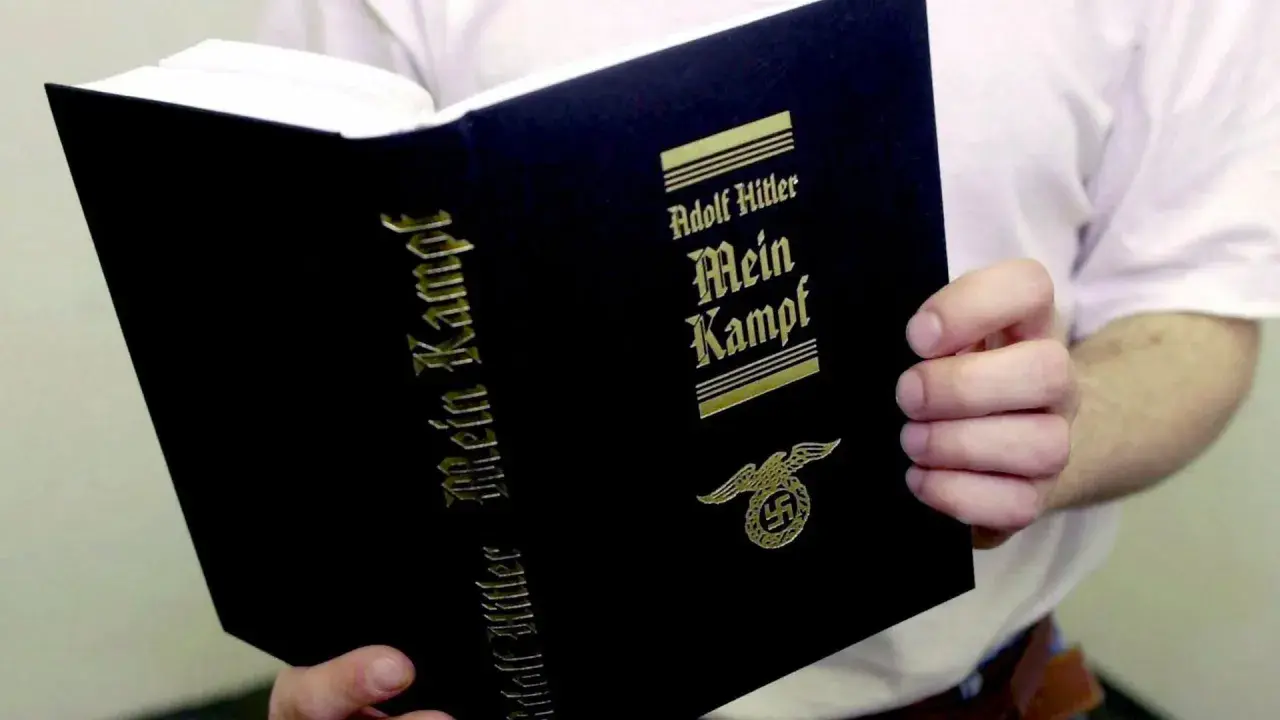 Eine Person hält ein Buch mit dem Titel "Mein Kampf" von Adolf Hitler. Das Buch hat einen schwarzen Einband mit goldener Schrift und einem Hakenkreuz-Adler-Symbol.
