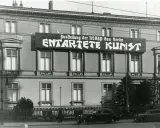 Große Banner verkünden die "Ausstellung der NSDAP Gau Berlin ENTTARTETE KUNST" an einem Gebäude. Autos und Fahrräder stehen davor.