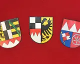 Drei Wappen auf rotem Grund: links ein Löwe, Mitte ein Adler, rechts ein Fähnlein und ein Rad. Sie repräsentieren bayerische Regierungsbezirke.