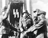 Mehrere SS-Soldaten stehen vor einer Fahne mit SS-Runen. Einer hebt den Arm zum Gruß.