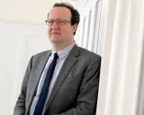 Hans Ottomeyer, ein Mann mit Brille im grauen Anzug, lehnt an einer weißen Wand.