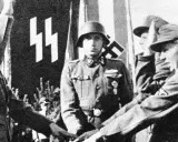 Ein SS-Offizier im Helm steht vor einer Fahne mit SS-Runen. Mehrere junge Männer in Uniformen sind zu sehen.
