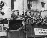 Männer in Uniformen, die an den Hitler-Putsch erinnern, stehen auf einem Lastwagen mit einer Fahne.