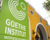 Grünes Schild mit weißem Logo und Schriftzug "GOETHE INSTITUT" vor einem gelben Gebäude.