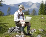 Mann in Lederhose und Hut arbeitet auf einem Laptop in den Bergen.