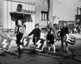Jungen in Uniformen tragen Fahnen mit Hakenkreuzen und Trommeln. Ein junger Mann, der an Hitler erinnert, marschiert mit ihnen.