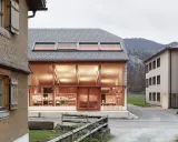 Neues Bauen: Moderne Architektur trifft auf traditionelle Schindelfassade. Ein hell erleuchtetes Glasgebäude mit Ausstellungsstücken steht neben einem Holzhaus.