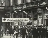 Menschenmenge vor einem Gebäude mit der Aufschrift "Berliner Schloßbräu-Quelle". Ein Banner verkündet "Deutsche Arbeiter! Wählt Liste 9". Hakenkreuzfahnen sind sichtbar, was auf die Zeit vor dem NSDAP Verbot hinweist.