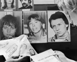 Zwei Personen betrachten Fahndungsfotos von Mitgliedern der RAF 2. Generation. Ein Plakat mit einer Belohnung von 300.000 DM liegt auf dem Tisch.
