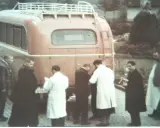 Männer in weißen Kitteln und dunklen Mänteln stehen vor einem roten Bus. Ein Kennzeichen ist sichtbar. Die Szene könnte mit der NS-Euthanasie in Verbindung stehen.