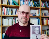 Mann mit Brille hält Foto eines Mannes in Uniform. Im Hintergrund Bücherregale. Das Foto zeigt einen Mann, der der SS Schutzstaffel angehörte.
