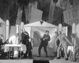 Szene einer Theateraufführung mit expressionistischem Bühnenbild. Menschen agieren auf der Bühne, als würden sie **trommeln in der Nacht**.