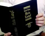 Eine Person hält ein Buch mit dem Titel "Mein Kampf" von Adolf Hitler. Das Buch hat einen schwarzen Einband mit goldener Schrift und einem Hakenkreuz-Adler-Symbol.