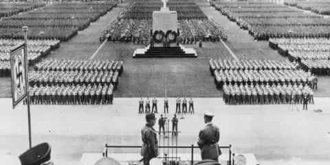 Massenveranstaltung in Nürnberg. Tausende marschieren, Fahnen wehen. Hitler ist anwesend.
