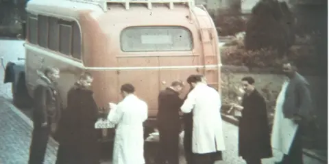 Männer in weißen Kitteln und dunklen Mänteln stehen vor einem roten Bus. Ein Kennzeichen ist sichtbar. Die Szene könnte mit der NS-Euthanasie in Verbindung stehen.
