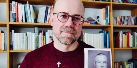 Mann mit Brille hält Foto eines Mannes in Uniform. Im Hintergrund Bücherregale. Das Foto zeigt einen Mann, der der SS Schutzstaffel angehörte.