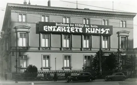 Große Banner verkünden die "Ausstellung der NSDAP Gau Berlin ENTTARTETE KUNST" an einem Gebäude. Autos und Fahrräder stehen davor.