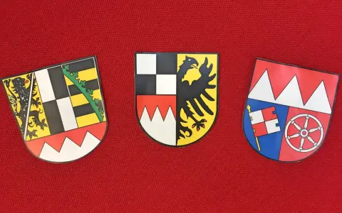 Drei Wappen auf rotem Grund: links ein Löwe, Mitte ein Adler, rechts ein Fähnlein und ein Rad. Sie repräsentieren bayerische Regierungsbezirke.