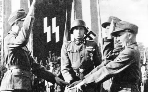 Mehrere SS-Soldaten stehen vor einer Fahne mit SS-Runen. Einer hebt den Arm zum Gruß.