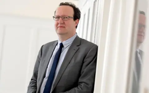 Hans Ottomeyer, ein Mann mit Brille im grauen Anzug, lehnt an einer weißen Wand.