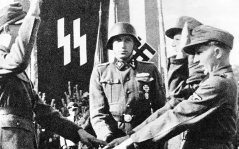 Ein SS-Offizier im Helm steht vor einer Fahne mit SS-Runen. Mehrere junge Männer in Uniformen sind zu sehen.
