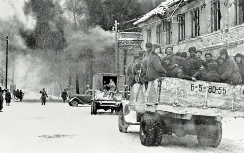 Soldaten der Roten Armee 1945 fahren in einem LKW durch eine verschneite Stadt. Rauch steigt aus einem zerstörten Gebäude auf.