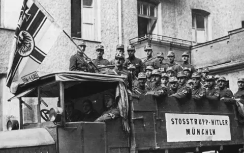 Männer in Uniformen, die an den Hitler-Putsch erinnern, stehen auf einem Lastwagen mit einer Fahne.
